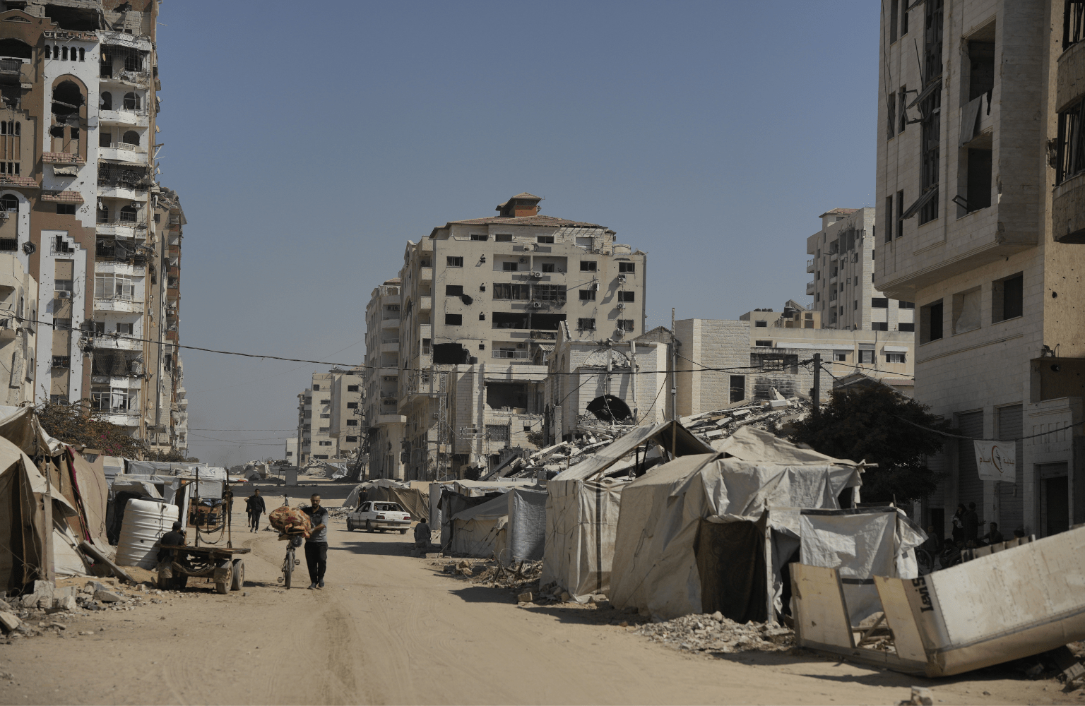 A street in Gaza lined with severely damaged and partially collapsed multi-story buildings, with makeshift tents set up