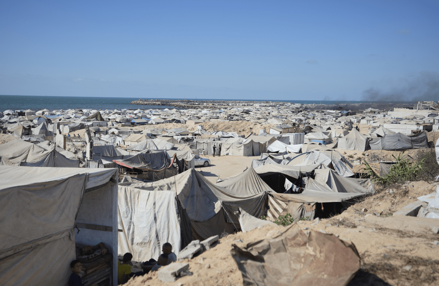 A wide photograph of a sprawling and crowded camp of makeshift tents for displaced people on a sandy coast in Gaza